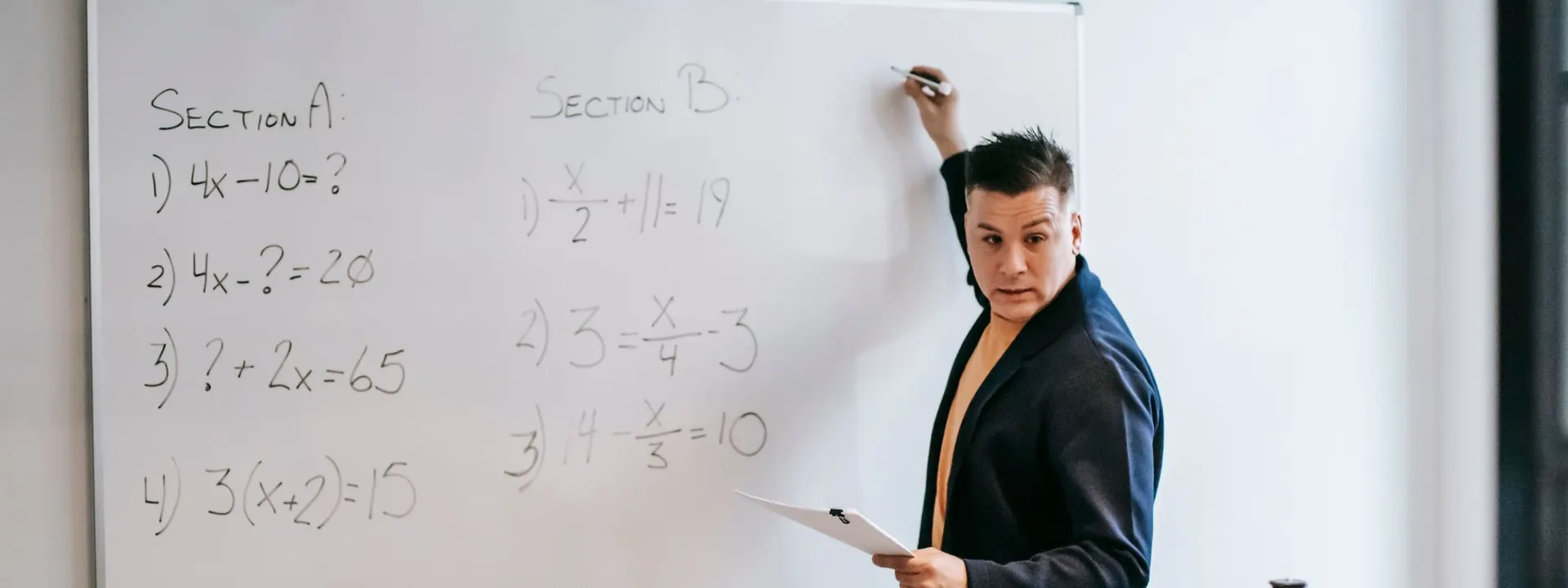 A whiteboard of worked algebra questions, with a tutor writing out the next step during a Mathematics class